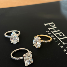 Load image into Gallery viewer, Three rings displayed on a black jewelry box: a pear-cut diamond ring in silver, an oval-cut diamond ring in yellow gold, and an emerald-cut diamond ring in silver. The brand name 'PHELANS JEWELLERS' is partially visible on the box.