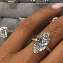 Load image into Gallery viewer, A close-up of a hand wearing a yellow gold ring with a large marquise-cut clear gemstone, with other rings displayed in the background on a gray velvet ring box.