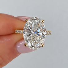 Load image into Gallery viewer, An oval diamond ring with a yellow gold band, held between fingers against a light gray background.