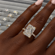 Load image into Gallery viewer, An emerald-cut diamond ring with a yellow gold band and prongs, shown worn on a person's finger. The ring features a brilliant clear center stone with smaller accent diamonds along the band, set against a blurred gray jewelry display background.