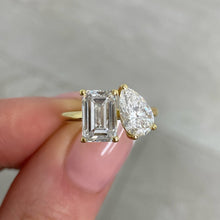 Load image into Gallery viewer, A close-up of a hand holding a yellow gold ring with an emerald-cut diamond and a pear-cut diamond, set against a soft gray marble background.