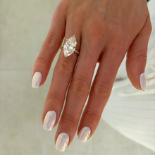 Load image into Gallery viewer, A close-up of a hand wearing a gold ring with a large marquise-cut clear gemstone. The nails are manicured in a pale iridescent pink color, and the background is a soft, light beige surface.

