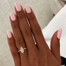 Load image into Gallery viewer, A close-up of a hand with long, glossy light pink manicured nails wearing a delicate gold ring with a marquise-cut clear gemstone. The background shows a light-colored tiled floor and a white ribbed fabric surface.
