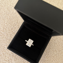 Load image into Gallery viewer, A rectangular diamond engagement ring in a silver setting, displayed inside an open black presentation box on a beige carpeted floor.
