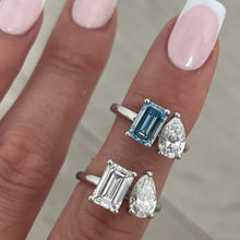 Load image into Gallery viewer, A close-up of a hand with a French manicure wearing two platinum rings, one featuring an emerald-cut clear stone and a pear-cut clear stone, vs one featuring an emerald-cut blue stone and a pear-cut clear stone.
